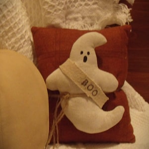 Op de afbeelding: Een wit spook kussen met een bruin lint waarop "BOO" staat. Het kussen ligt op een rood en wit kussen.