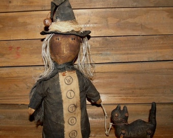 May include: A handmade rag doll witch wearing a black dress and a pointed black hat with a brown brim. The doll is holding a string attached to a black cat with a white face. The doll has a brown face and white hair. The doll is standing on a wooden surface. The doll's dress has the word "WITCH" written on it.