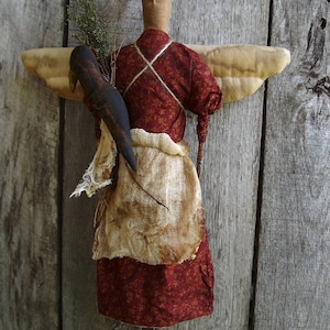 Puede incluir: Un ángel de trapo hecho a mano con la cara marrón, vestido rojo y un pájaro marrón en sus brazos. El ángel tiene un halo y está de pie contra un fondo de madera.