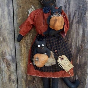 May include: Black cat doll wearing a red and black plaid dress and holding two smaller black cat dolls, each holding a small orange pumpkin. The larger doll has a tag that reads "Day At The Patch".