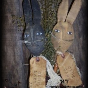 May include: Two primitive bunny dolls with fabric bodies and painted faces. The bunnies are holding tags that say "Olde Hare" and "Olde Fare".