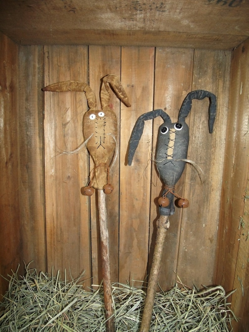 May include: Two handmade primitive bunny dolls with bells on sticks. The bunnies are made of fabric and have button eyes. One bunny is brown and the other is black. The bunnies are standing in a wooden crate filled with hay.