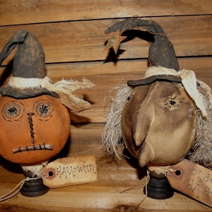 May include: Two handmade Halloween witch dolls with burlap fabric and stitched features. One doll is a pumpkin with a black hat and the other is a witch with a black hat and long white hair. The dolls are on wooden bases and have tags that say "Halloween" and "Old Hag".