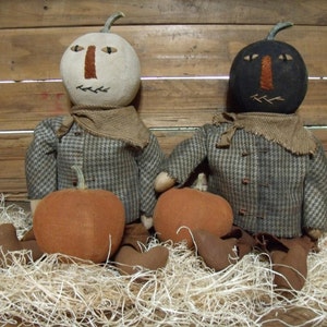 May include: Two handmade pumpkin people dolls with brown and white checked shirts and brown scarves. Each doll is holding a small orange pumpkin.