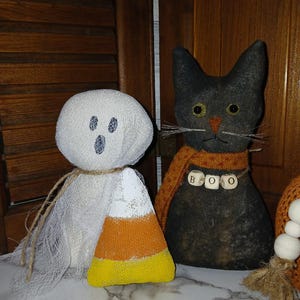 May include: Handmade Halloween decorations featuring a ghost, a black cat, and a pumpkin. The ghost is white with blue eyes and a candy corn base. The black cat wears a scarf with "BOO" spelled out. The pumpkin is orange.
