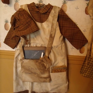 Puede incluir: Un vestido marrón y blanco con un corpiño de cuadros marrones y un delantal blanco. El vestido tiene un ribete de cuadros marrones y un sombrero de cuadros marrones. El vestido tiene un bolsillo con el texto "Emma Grade 3 - 1805" y otro bolsillo con el texto "Reader".