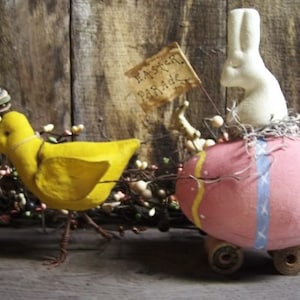 May include: A yellow chick with a blue and green hat pulls a pink and white Easter egg on wheels. The egg has a white bunny on top and a sign that says "Easter Parade".