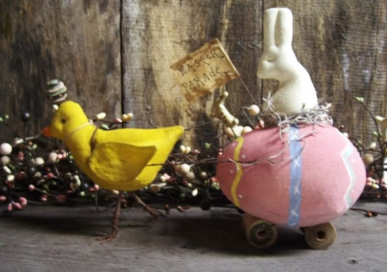 May include: A yellow chick with a blue and green hat pulls a pink and white Easter egg on wheels. The egg has a white bunny on top and a sign that says "Easter Parade".