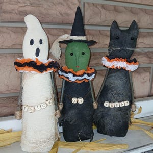 Pode incluir: Três figuras de Halloween feitas à mão: um fantasma, uma bruxa e um gato preto. O fantasma é branco com olhos e boca pretos, a bruxa tem um rosto verde e um chapéu preto, e o gato tem bigodes. Cada figura tem uma gola franzida e uma faixa de palavras de madeira.