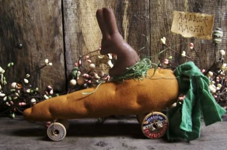 Puede incluir: Un conejo marr&oacute;n se sienta en la parte superior de un coche con forma de zanahoria con tela verde drapeada sobre la parte posterior. El coche tiene dos ruedas con radios y las palabras "Rabbit Racer" est&aacute;n escritas en un cartel detr&aacute;s del coche.