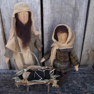 May include: A handmade nativity scene with three rag dolls. The figures are dressed in brown and beige fabric. The baby doll is lying in a manger made of wood and straw.