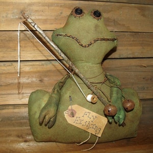 May include: A handcrafted, green fabric frog doll with button eyes and a stitched mouth. It holds a fishing rod with a hook and a tag that reads "FLY FISHING". Two small bells hang from its arm.