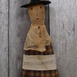 May include: A handmade rag doll witch with a black pointed hat, a brown and tan plaid skirt, and a burlap apron with the text "BROOMHILDA" embroidered on it. The doll is standing against a weathered wooden background.