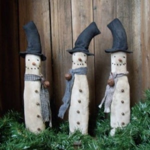 Könnte beinhalten: Drei weiße Stoff-Schneemannfiguren mit schwarzen Hüten und blauen Schals. Jeder Schneemann hat eine braune Knopfnase und braune Knöpfe für die Augen. Die Schneemänner stehen vor einem hölzernen Hintergrund mit grünem Kranz.