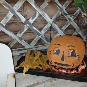 May include: A hand-painted orange pumpkin-shaped container with a smiling face, black handle, and a ruffled collar. It sits on a wooden tray with dried corn husks, perfect for Halloween decor.