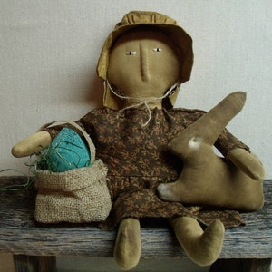 May include: A handmade rag doll dressed in a brown floral print dress and a brown bonnet holds a burlap bag with a blue and green egg inside. The doll is sitting on a wooden bench with a brown stuffed bunny beside it.