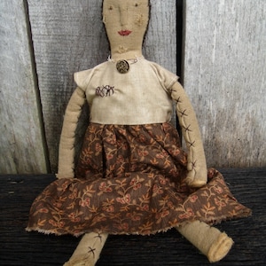 May include: A handmade rag doll with a brown and tan floral print dress. The doll has a stitched face with brown eyes and red lips. The doll is sitting on a wooden surface.
