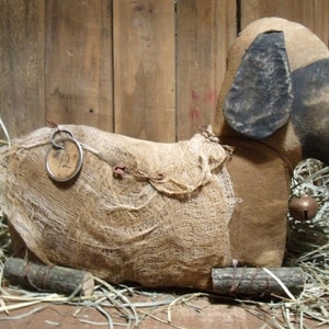 Può includere: Un cane di stoffa in stile primitivo marrone e beige con naso e orecchie in stoffa nera. Il cane indossa un sacco di juta e ha un cartellino di metallo con il numero "12" sopra. Il cane è seduto su un letto di paglia.