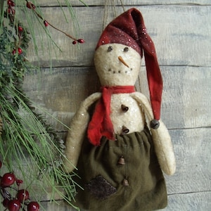 May include: A handmade snowman ornament with a red hat, red scarf, and brown fabric skirt. The snowman has a white face with black eyes and a carrot nose. The ornament is decorated with bells and is hanging from a string.