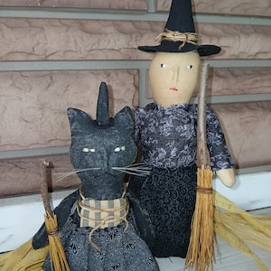 Puede incluir: Dos muñecas de Halloween hechas a mano: un gato negro con un sombrero de bruja y una muñeca de bruja. El gato tiene una escoba y una bufanda a cuadros. La muñeca de bruja lleva un sombrero negro y lleva una escoba. Ambas muñecas están vestidas con tela negra.
