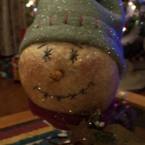 May include: A whimsical snowman ornament with a tan face, embroidered eyes and smile, and a carrot nose. It wears a gray hat with red stitching and a red scarf. The ornament is decorated with glitter and is likely for Christmas.