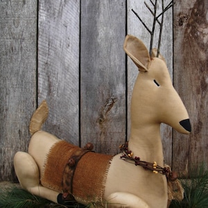 以下が含まれることがあります： 茶色の布地でできた手作りのかわいい鹿の置物。鹿は枝の角と、首に茶色と赤のベリーのガーランドをつけています。鹿は松葉のベッドの上に座っています。