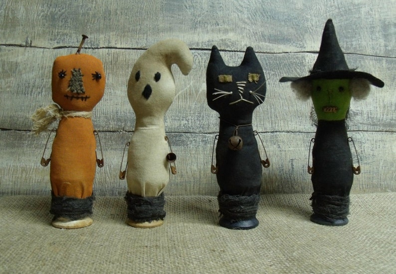 May include: Four handmade Halloween-themed dolls. A pumpkin, ghost, black cat, and witch. Each doll is made of fabric and has safety pins for arms and legs. The dolls are standing on a hessian surface.