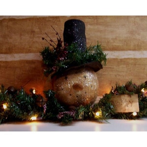 Pode incluir: Uma figura de boneco de neve com um chapéu-coco preto e um rosto brilhante, cercada por uma guirlanda verde com luzes e detalhes marrons. A guirlanda tem uma etiqueta de papel marrom com a palavra "Cozy" escrita nela.