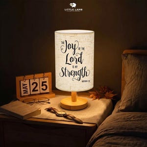 May include: A cylindrical table lamp with a light-colored wooden base and a textured white fabric shade. The shade features black cursive text that reads The Joy of the Lord is My Strength Nehemiah 8:10. The lamp emits a warm, soft glow.