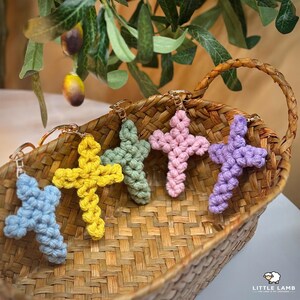 Puede incluir: Cinco llaveros de cruz de macramé en una cesta tejida. Los llaveros son de color azul, amarillo, verde, rosa y morado. Cada cruz tiene un llavero dorado. La cesta es marrón y tiene un asa.