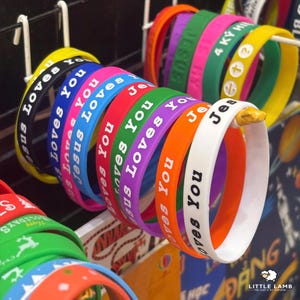 Könnte beinhalten: Eine Auswahl an bunten Armbändern aus Gummi mit dem weißen Schriftzug "Jesus Loves You". Die Armbänder sind in verschiedenen Farben erhältlich: Gelb, Schwarz, Blau, Rosa, Rot, Grün, Lila, Orange und Weiß.