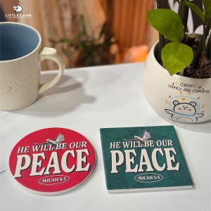 May include: A round red coaster and a square green coaster with the text "HE WILL BE OUR PEACE" and "MICAH 5:5". A white mug with a blue interior and a small white pot with a plant are also visible.