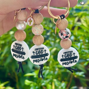 May include: Three keyrings with wooden beads, a cross charm, and a white disc with the text "With God All Things Are Possible." The beads include natural wood, a pearl, and patterned designs. Gold-coloured key rings.