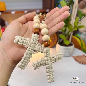 Puede incluir: Llavero de cruz de macramé con cuentas de madera. La cruz está hecha de cordón de macramé blanquecino, y el llavero presenta cuentas de madera natural y un anillo de metal dorado. El diseño es artesanal y tiene una estética rústica y bohemia.