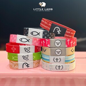 Pode incluir: Uma pilha de pulseiras de tecido coloridas com símbolos religiosos e texto. As pulseiras apresentam cruzes, peixes, pombas e ovelhas. O texto inclui "Little Lamb" e "The Lord is my Shepherd".