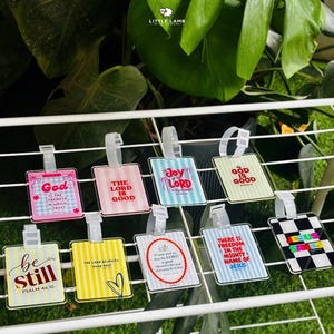 May include: A collection of ten rectangular luggage tags featuring various designs, including striped patterns in pink, yellow, and blue, a black and white checkerboard print, and religious text such as God is the strength, The Lord is good, and Be still.