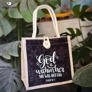 Könnte beinhalten: Eine kleine Tragetasche mit einer schwarzen Frontplatte mit floralem Muster und dem weißen Text "God is within her, she will not fall, Psalm 46:5." Die Tasche hat einen beigefarbenen Jute-Körper, cremefarbene Griffe und einen Knopfverschluss.