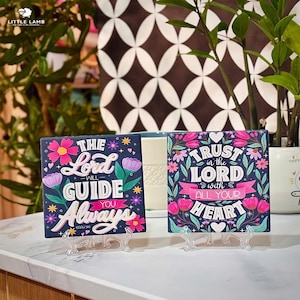 Può includere: Due piastrelle decorative quadrate con disegni floreali e citazioni ispiratrici. Una piastrella recita "The Lord Will Guide You Always" e l'altra "Trust in the Lord with All Your Heart". Entrambe sono blu navy con fiori colorati.