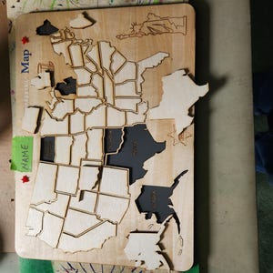 May include: Wooden United States map puzzle with state outlines and state names. The states of Alaska, Hawaii, and others are cut out in dark gray. The word "Name" is written on a green sticky note.