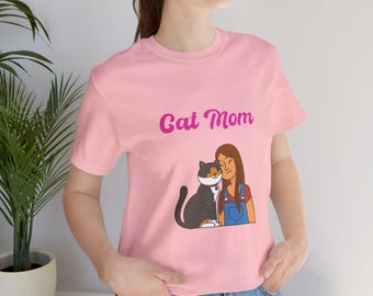 May include: A pink t-shirt with the text "Cat Mom" in pink lettering. A cartoon illustration of a woman with brown hair hugging a black and white cat is on the shirt.