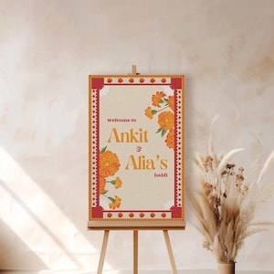 May include: Welcome to Ankit & Alia's Haldi sign with orange flowers on a brown background with a red border. The sign is on an easel.