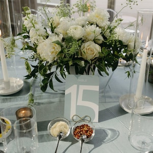 Puede incluir: Un centro de mesa floral blanco con vegetación se encuentra sobre un mantel azul claro. Una tarjeta de número de mesa con el número 15 y la palabra "TABLE" está delante de las flores. Dos cuencos pequeños con cucharas contienen condimentos blancos y rojos.