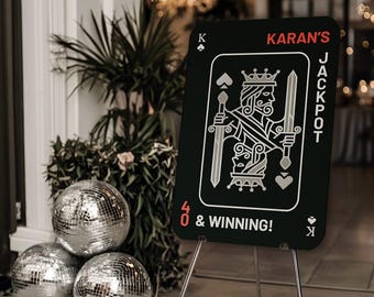 May include: A rectangular black sign shaped like a playing card on a metal tripod stand. The sign features a white king illustration, the text KARAN'S JACKPOT, and 40 & WINNING! in red and white. The design includes spade and heart symbols.