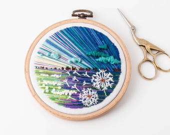 May include: An embroidered landscape scene in a wooden hoop. The artwork features a blue and green sky, fluffy clouds, and a field of dandelions. A pair of gold-colored scissors rests beside the hoop.