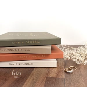 May include: A stack of four beige and brown hardcover journals with gold lettering on the spines. The top journal reads "Liam & Georgia", the second reads "Lucianna & Richard", the third reads "Nicholas & Veronica", and the bottom reads "David & Hannah". The journals are on a wooden surface with a small bouquet of white flowers and two gold rings.