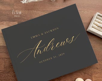 May include: A charcoal gray guest book with gold foil lettering. The text reads "Emma & Jackson Andrews" and "October 31, 2029".
