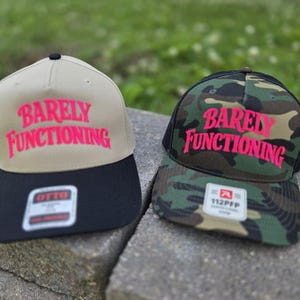 May include: Two baseball caps with the text BARELY FUNCTIONING in bright pink. The left cap is tan with a black brim and an OTTO sticker. The right cap features a green, brown, and black camouflage pattern with a black brim and a 112PFP sticker.