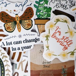 Pode incluir: Uma coleção de autocolantes decorativos com vários desenhos e texto. Um autocolante apresenta uma borboleta, outro diz "A lot can change in a year". Um terceiro autocolante diz "I'm still learning."