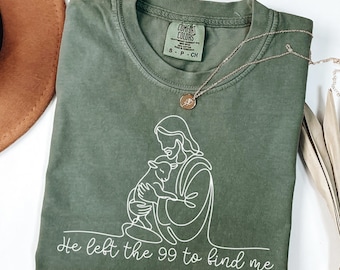 May include: A moss green t-shirt with a white line drawing of a figure holding a lamb. The text "He left the 99 to find me" is below the drawing. A gold necklace with a pendant is on the shirt.