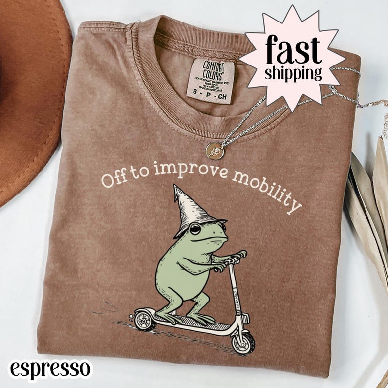 Espresso graphic shirt featuring an illustrated frog wearing a wizard hat riding a scooter with “Off to improve mobility” text, whimsical cartoon design